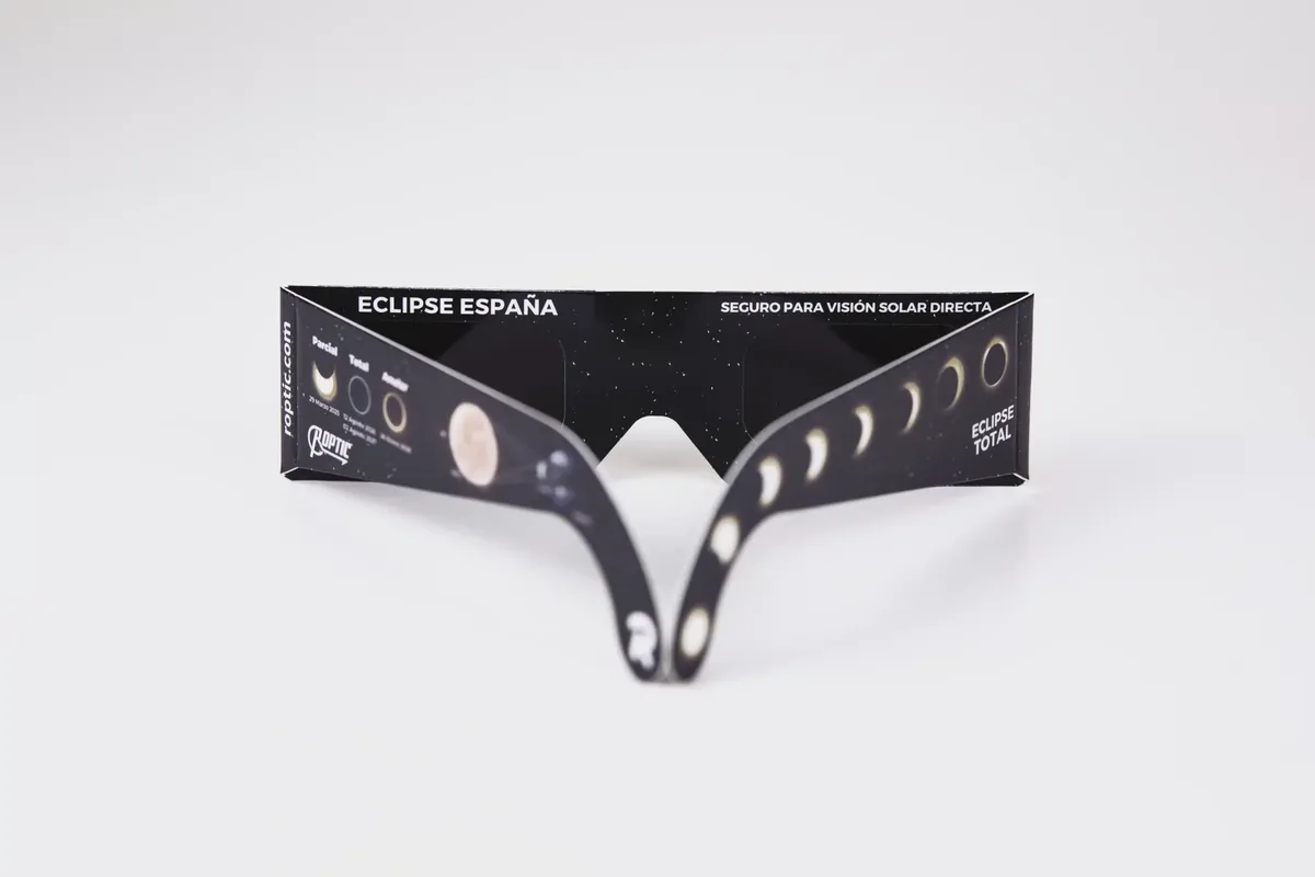 Gafas de eclipse ROPTIC de frente sobre fondo blanco, con el rótulo «Eclipse España · Seguro para visión solar directa» y la secuencia del eclipse impresa en la patilla