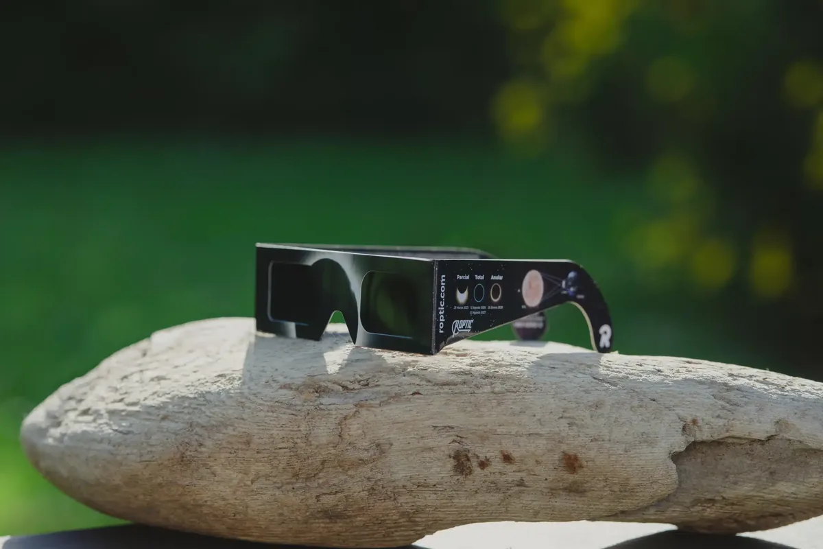 Gafas de eclipse ROPTIC apoyadas en una piedra al aire libre; en la patilla, los eclipses parcial, total y anular con sus fechas de 2025 a 2028